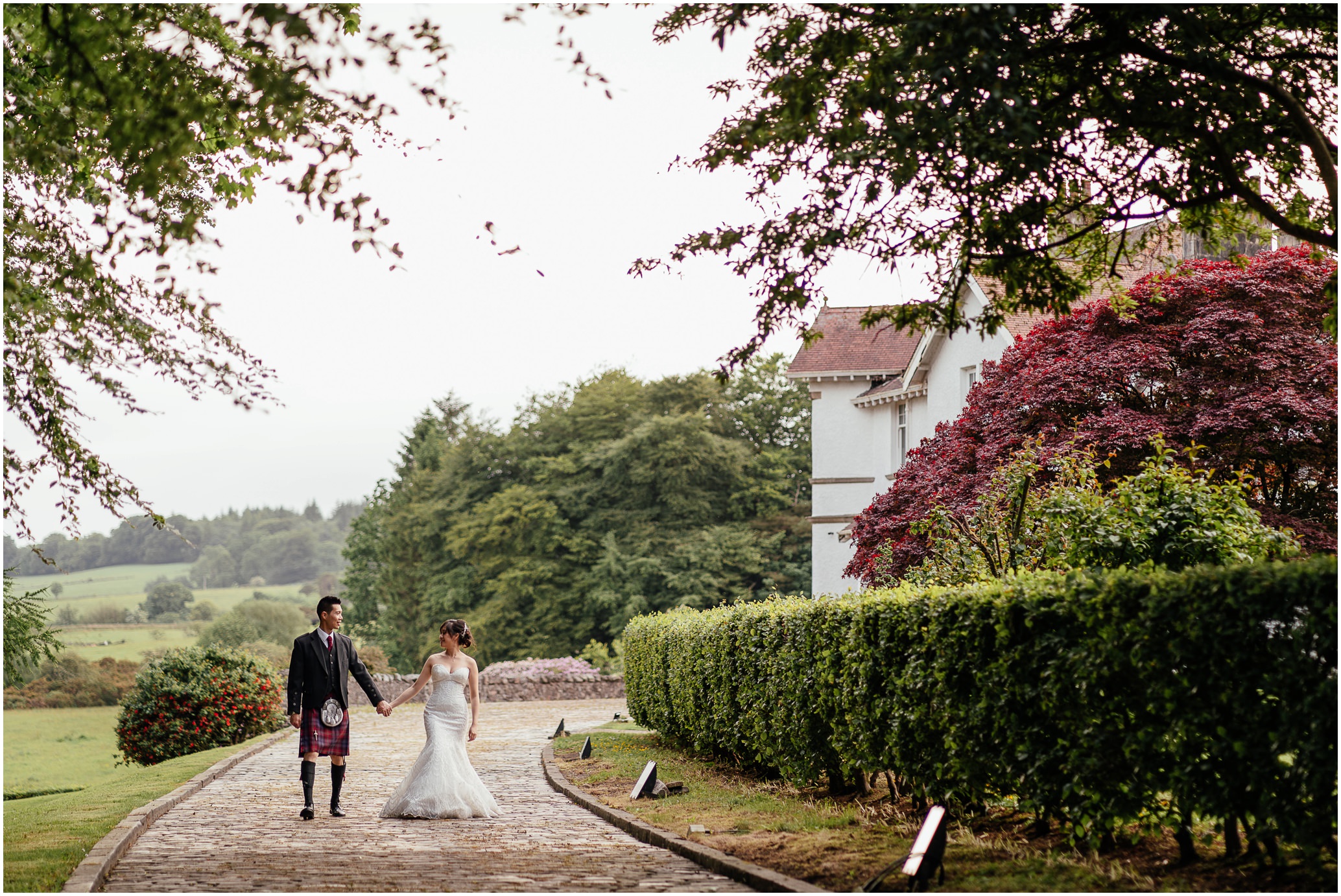 bride wearing white dress and groom in kilt outfit walking hand in hand down cobbled entrance to the estate green hedges trees in foreground and background and estate house post wedding photoshoot glasgow fotomaki photography