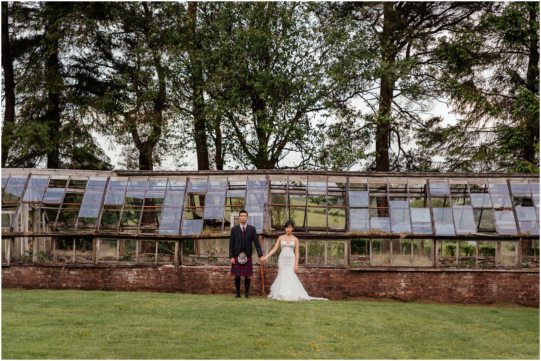 bride and groom hand in hand standing on grass lawn in front of rustic greenhouse in walled garden big trees in background post wedding photoshoot glasgow fotomaki photography
