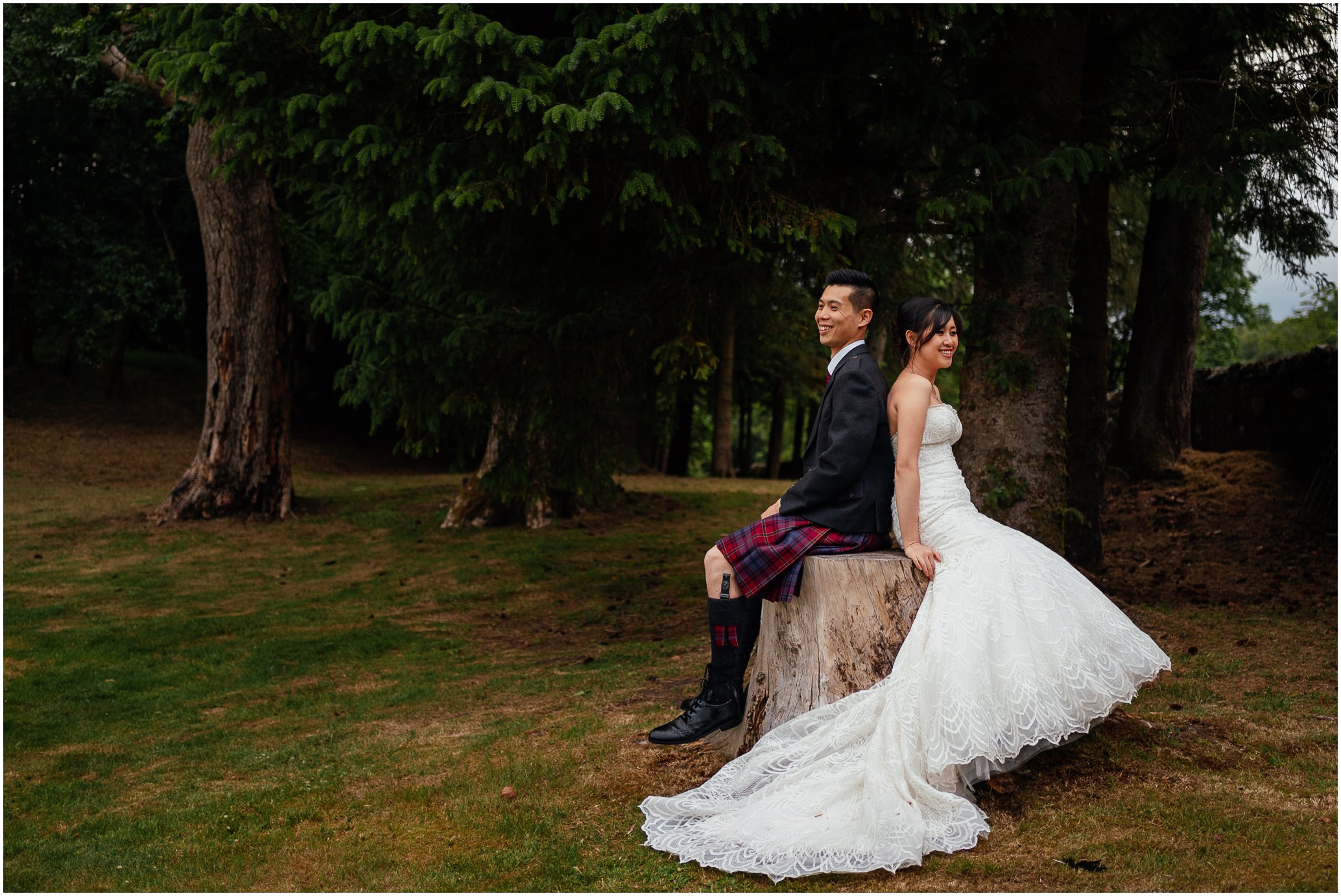 bride in white dress and groom in kilt outfit sitting back to back on tree stump surrounded by grass lawn and big trees in background post wedding photoshoot glasgow fotomaki photography