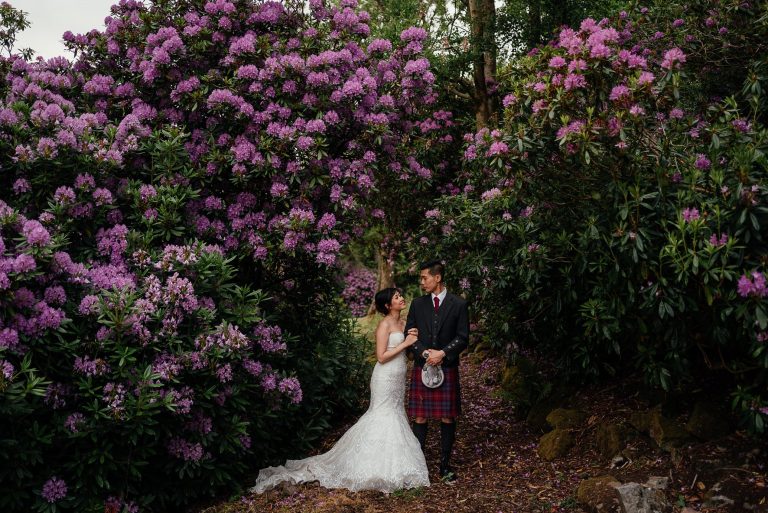 bride and groom with linked arms looking lovingly into teach other eyes under an arch of green bushes with purple flowers post wedding photoshoot glasgow fotomaki photography