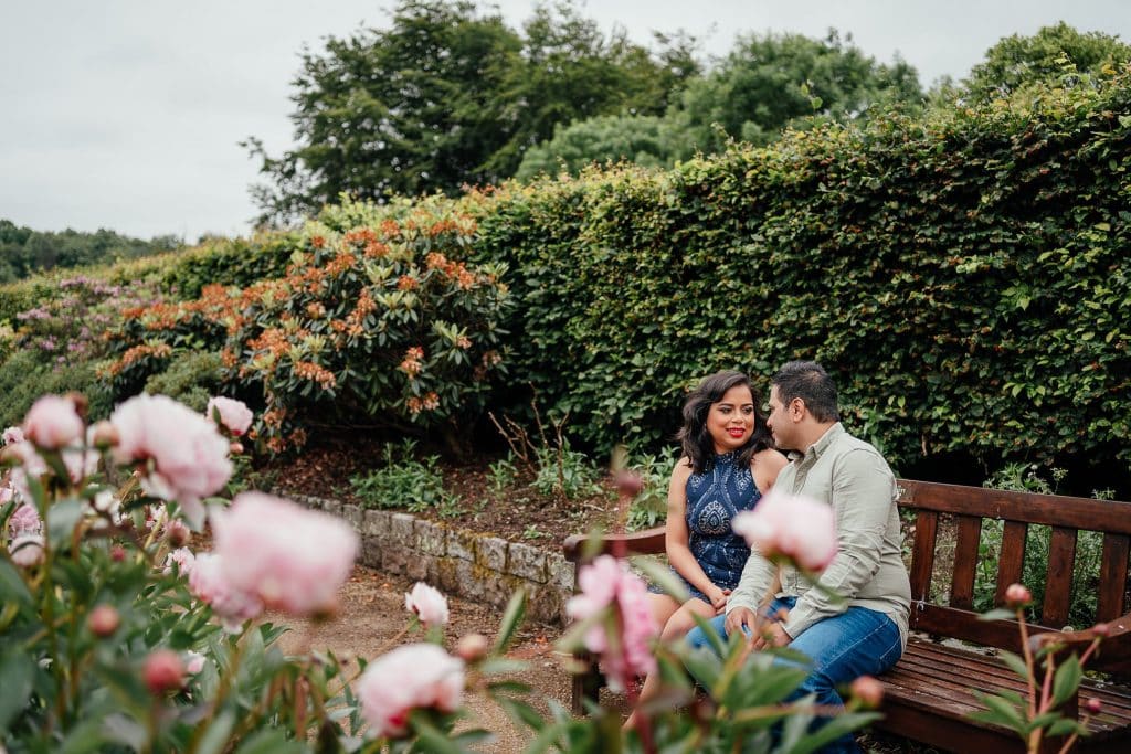 couple sit on bench surrounded by flowers in pollok park walled garden on wedding anniversary photoshoot glasgow