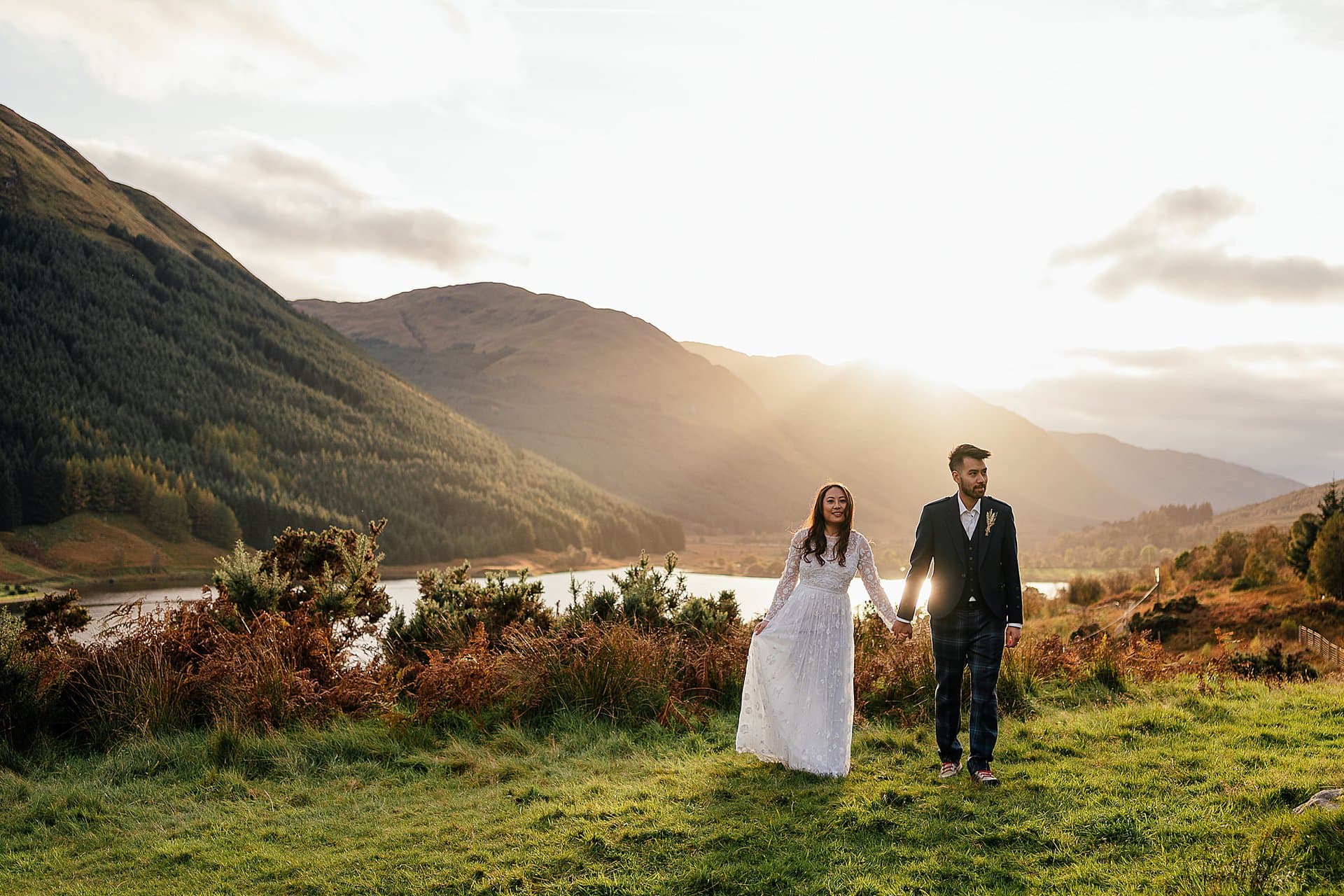 bride in white dress and groom wearing suit hand in hand on grass banking above loch with sun setting over green mountains in background monachyle mhor wedding photoshoot fotomaki photogrphy