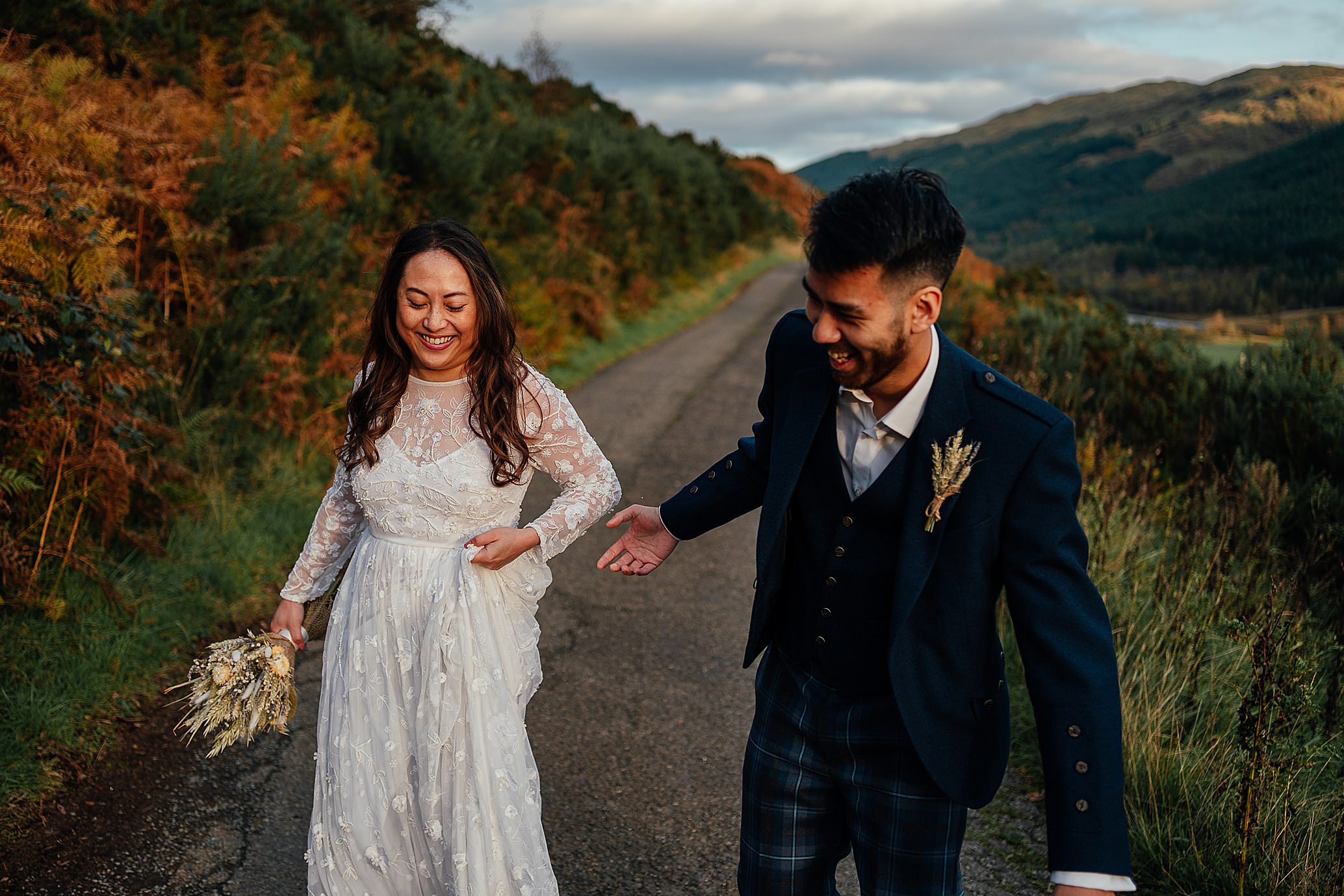 bride in stunning white dress and groom in suit jacket with kilt trousers walking and laughing on country road green banking and mountains in background monachyle mhor wedding photoshoot fotomaki photography