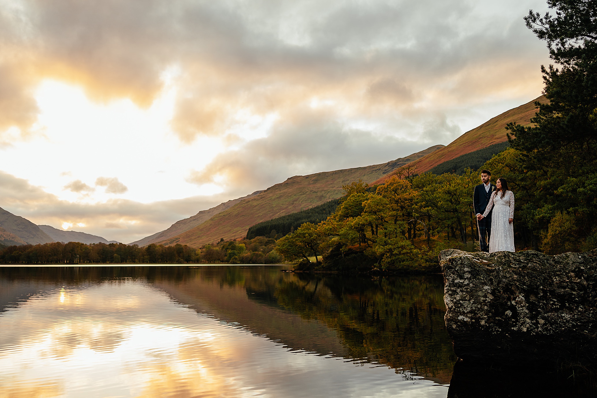 bride and groom standing on rock at side of loch holding hands as the sun sets green mountains in background monachyle mhor wedding photoshoot fotomaki photography