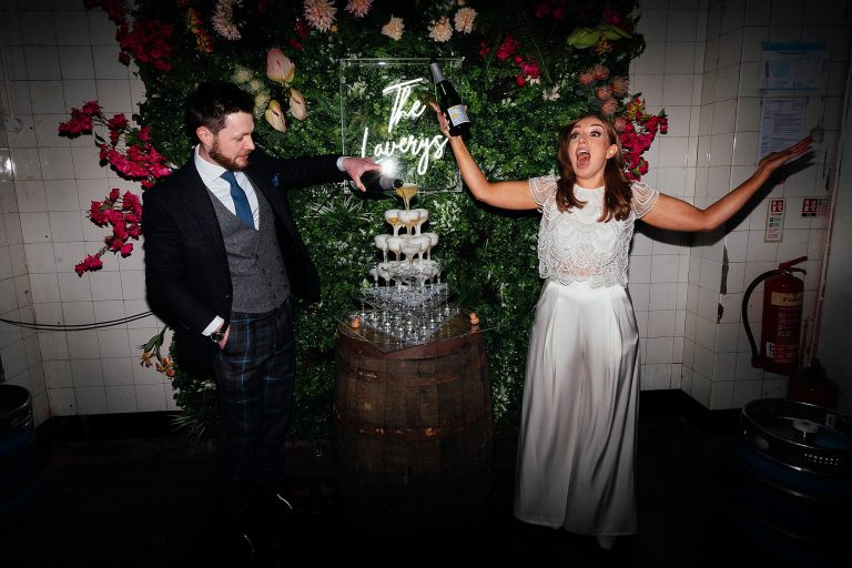 bride holding a champagne bottle with her hands in the air as groom fills champagne tower with plant wall in background biscuit factory edinburgh wedding fotomaki photography