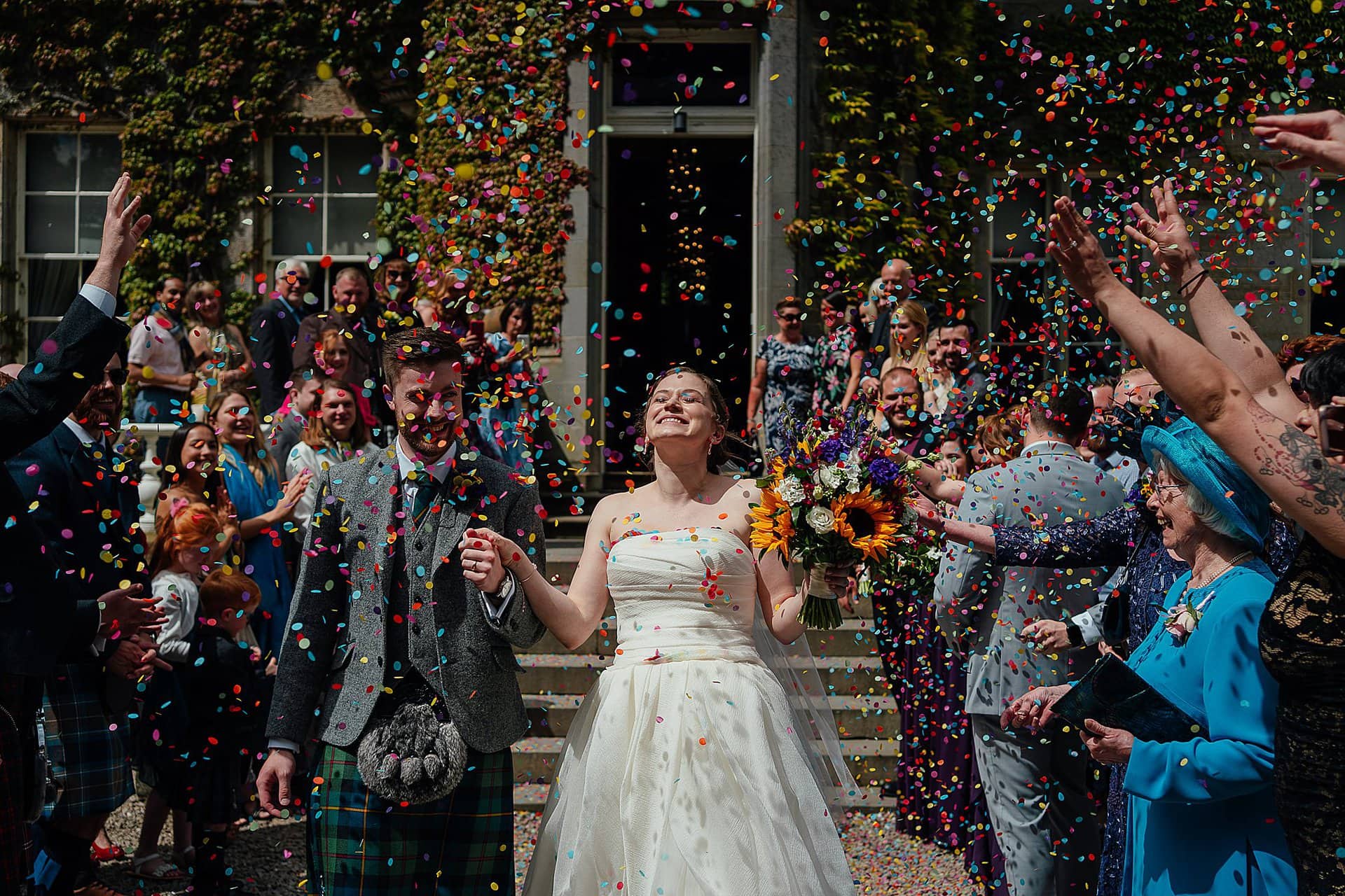 bride and groom holding hand and smiling as they walk through guests throwing confetti carlowrie castle wedding edinburgh colourful wedding photography scotland