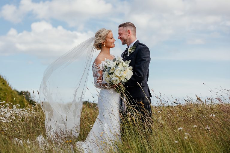 bride and groom looking into each others eyes brides veil blowing in the wind as they stand in grass field with blue skies in background falside mill wedding unique venues scotland