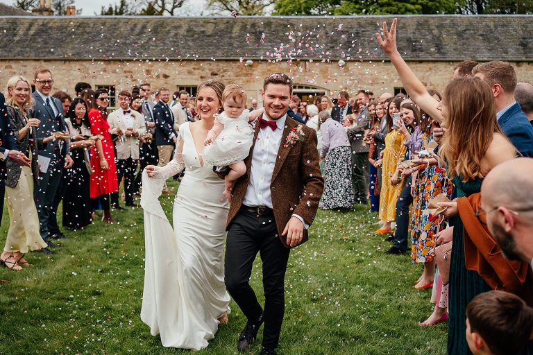 bride and groom carrying their baby as wedding guests shower them with confetti on grass lawn kirknewton stables wedding edinburgh