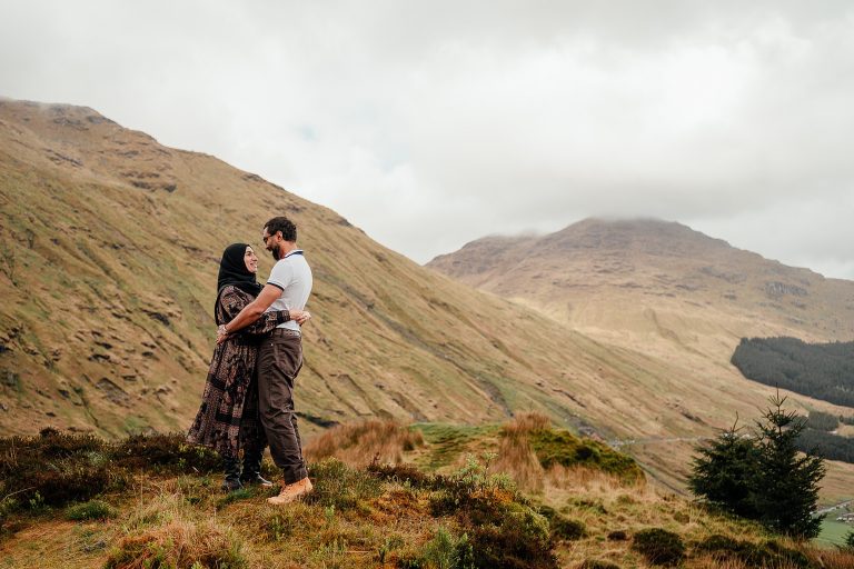 couple standing on grass clearing on hill with green mountains in background loch lomond photoshoot glasgow couples photos glasgow