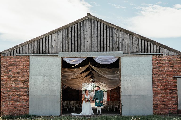 bride and groom hold hands at entrance of barn, the barn is set up with white drapes hanging from ceiling farm wedding scotland marquee wedding