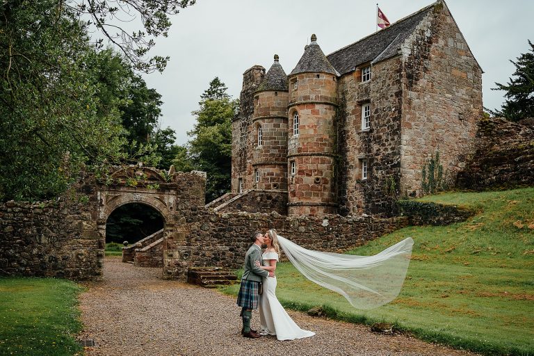 bride veil blows in the wind as she kisses the groom with the castle in background scottish rowallan castle wedding
