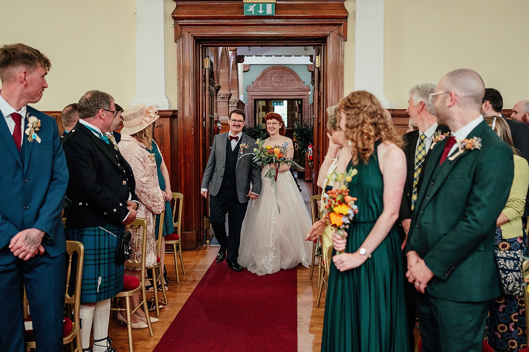 bride and groom walk hand in hand down the aisle as guests look on DIY pollokshields burgh hall wedding scotland
