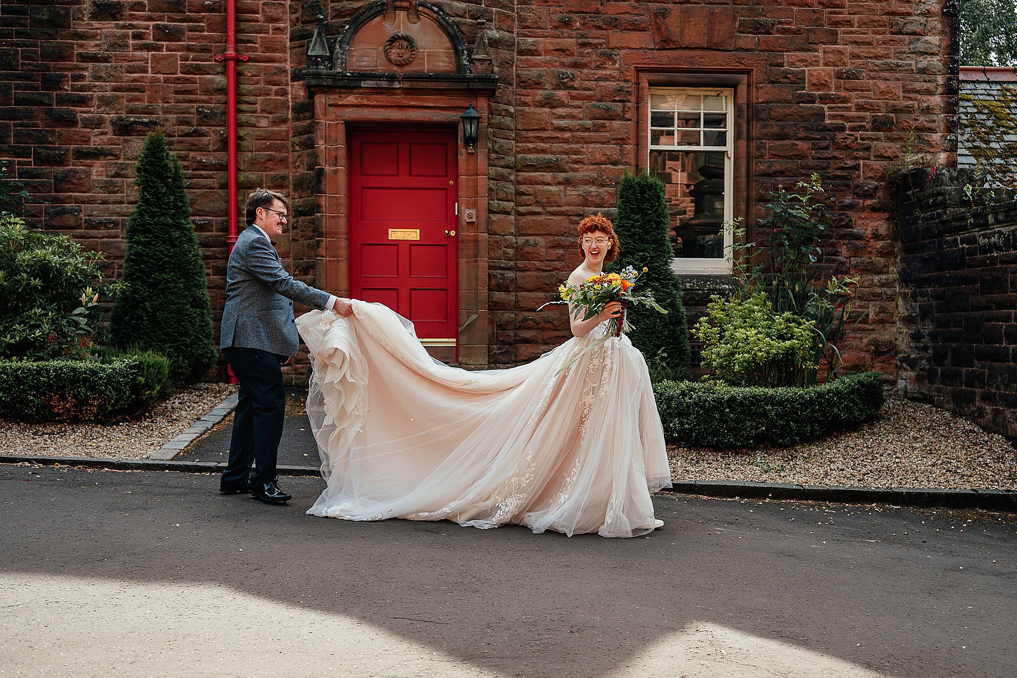 bride laughing as groom carries back of her dress whilst they walk through venue grounds quirky pollokshields burgh hall wedding glasgow