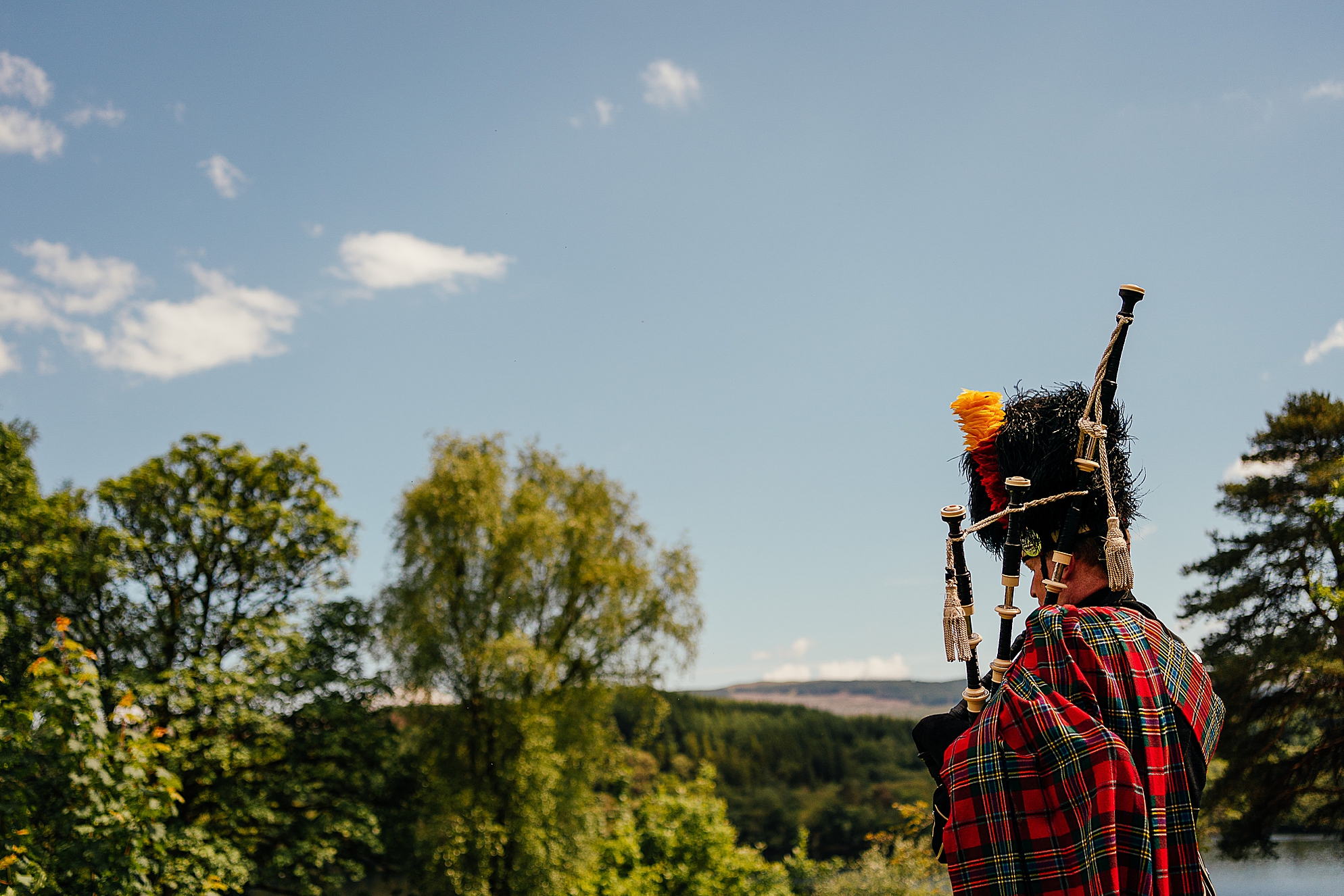 Scottish piper in highland dress tartan standing outdoors on hillside above loch awe for a kilchurn castle wedding in scotland