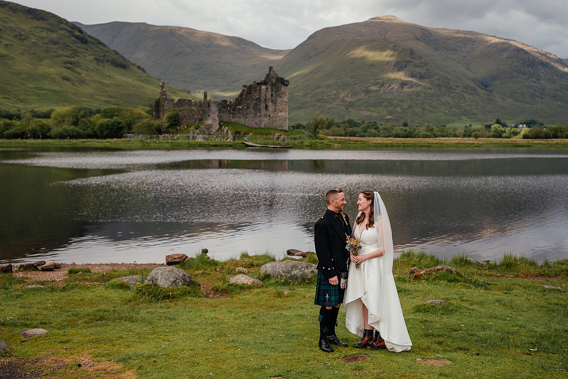 bride and groom smiling at each other in front of view of kilchurn castle for their micro wedding outdoors on loch awe