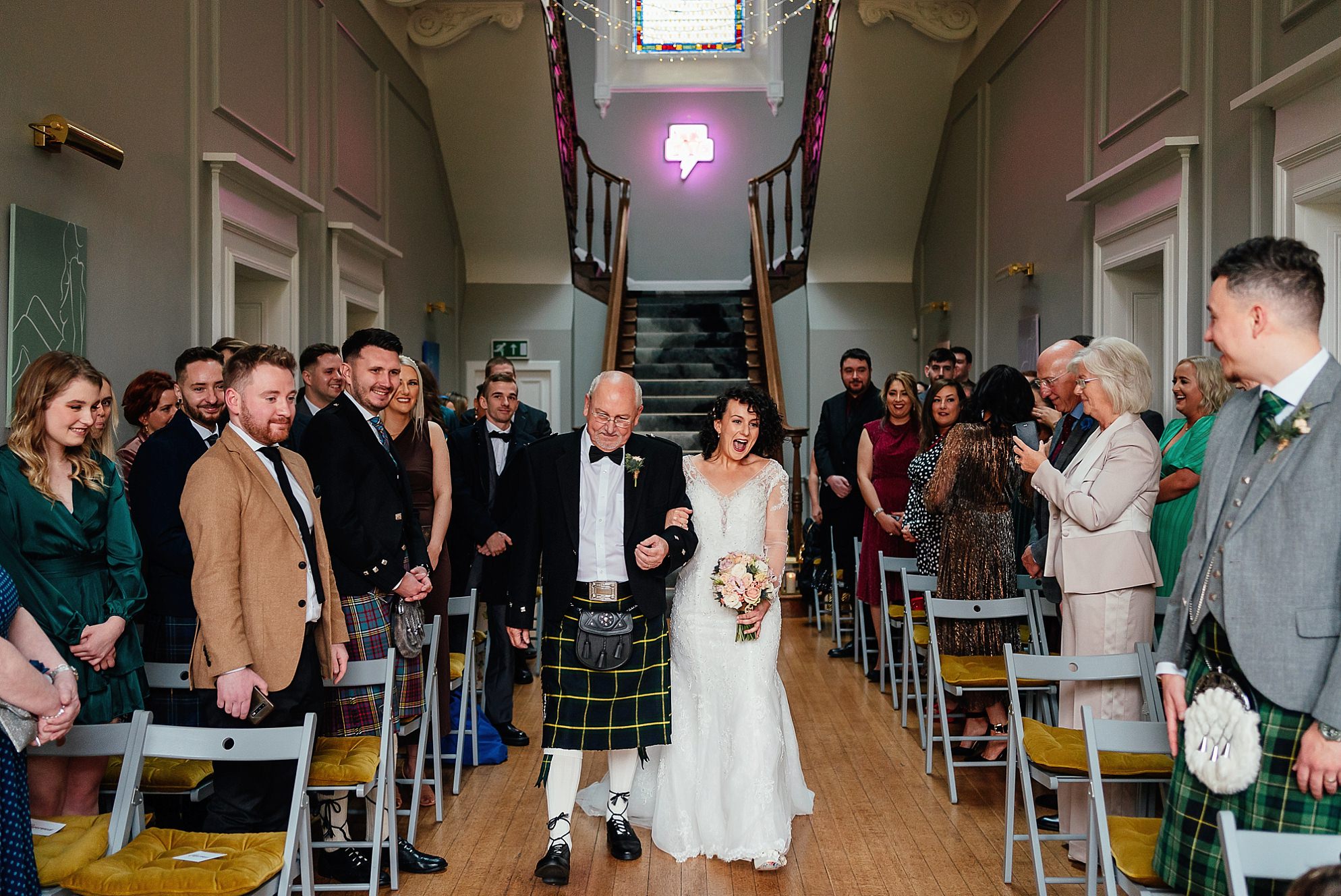 Interior inside Netherbyres House Wedding near Eyemouth in Scotland Guests family and friends stand up from their sea-grey folding chairs with colourful mustard cushions to welcome the bride in a lace gown as she walks down the aisle accompanied by an older gentleman in a kilt Bright pink fun neon sign and fairy lights light up the carved wooden victorian grand staircase in the ornate grand hall