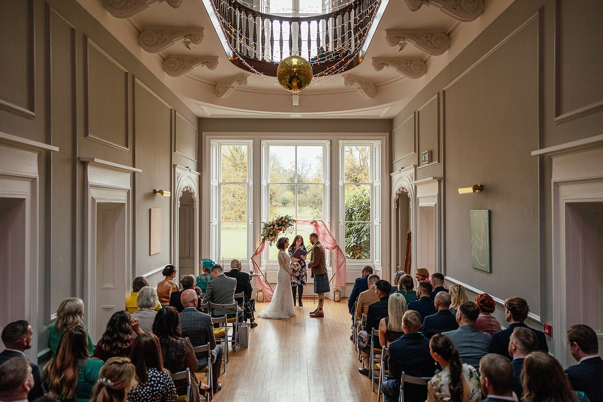 Interior inside grand hall DIY wedding venue at Netherbyres House near Eyemouth in Scotland Fiona Flanagan Humanist performs the ceremony with bride and groom at a colourful geometric wedding arch decked with flowers by Occasions of Eyemouth Florist gold mirrorball white fairy lights and abstract modern art mix perfectly with the period features of the traditional victorian stately house rear view of guests as they watch seated in sea-grey folding wooden seats with mustard cushions traditional victorian cornicing sash windows and modern art