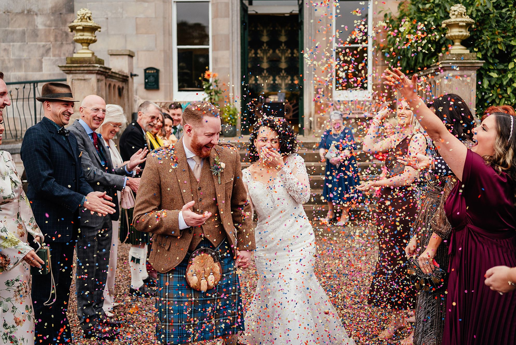 outside exterior view of Netherbyres House wedding in Eyemouth Scotland on an Autumn day A laughing fun-filled bride and groom walk through a crowd of bright and colourful smiling guests throwing confetti and delighting as the bride laughs at having lots of confetti in her mouth The guests are in a red gravel filled courtyard by traditional Victorian era red sandstone steps at an ornate porch flanked with arched stone banisters topped with gold urn-shaped sculptures  and surrounded by trees