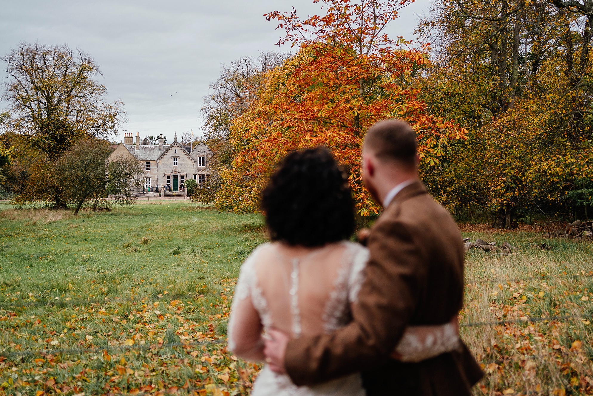 outside exterior view on the grounds of Netherbyres House in Eyemouth Scotland autumn wedding A rear view of the bride and groom as they look towards the rustic and characterful large Victorian stately home surrounded by mature trees and a lawn covered by colourful autumn leaves with autumnal tones of red yellow green and brown