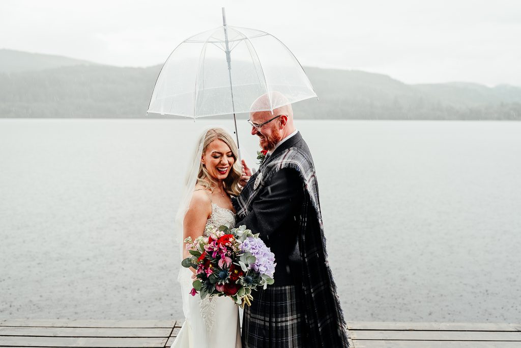 bride and groom standing together in the rain underneath an umbrella laughing at their loch venachar wedding