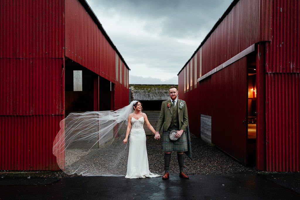 bride and groom standing between two red dutch barns and smiling at dalduff farm wedding venue