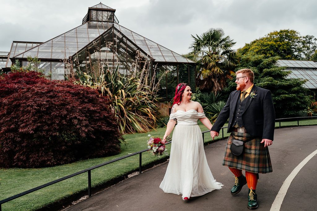 bride with red hair and groom with a kilt and orange socks walking hand in hand through the glasgow botanic gardens with a greenhouse in the background for their glasgow botanic gardens wedding portraits