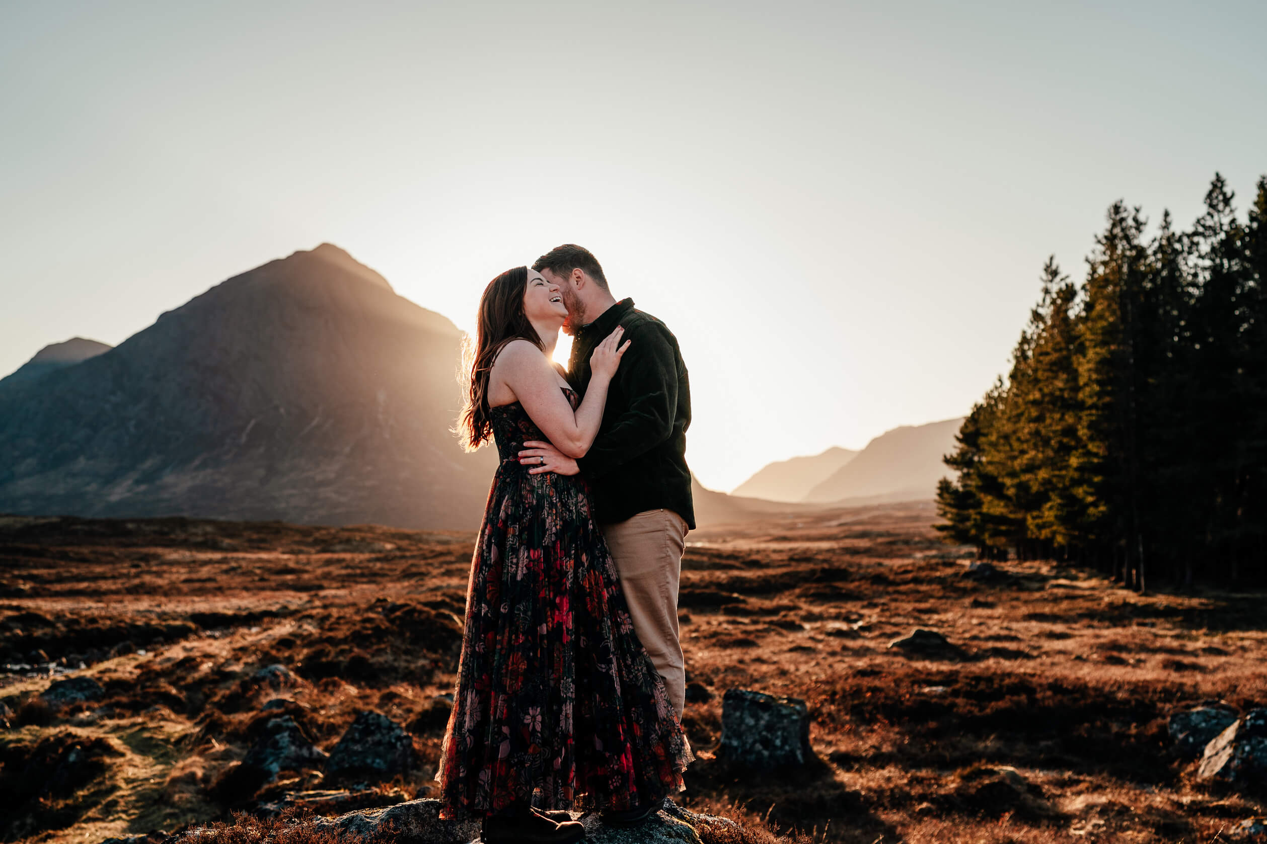 a couple stand in front of a mountain in glencoe as the sun sets behind them in scotland