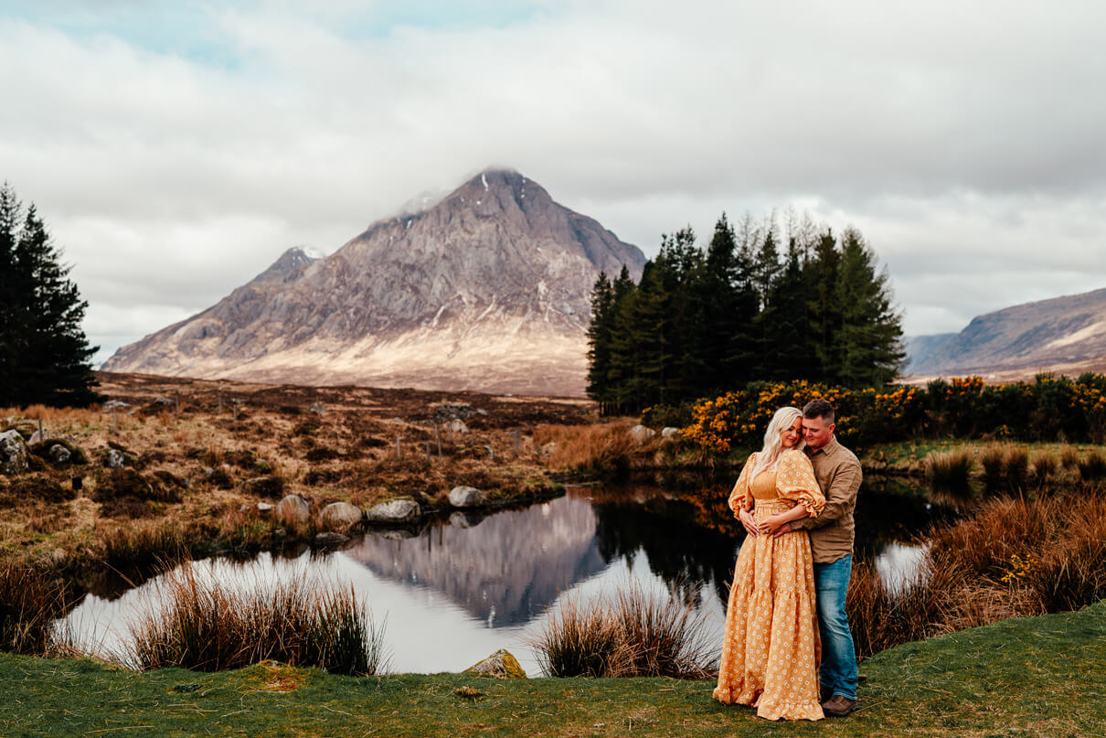 couple in yellow stand in front of mountain in glencoe that's reflected in the water