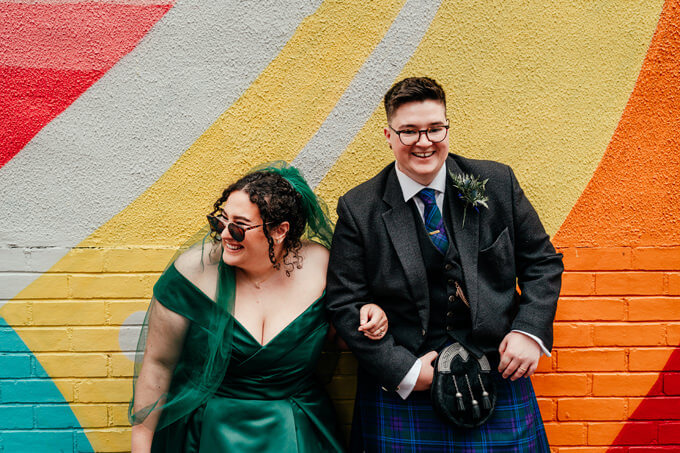 bride and trans groom laugh on their wedding day in front of colourful wall in glasgow